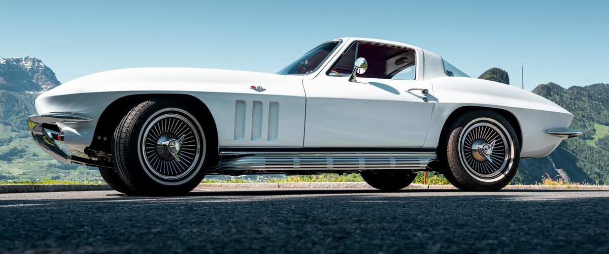 Low side view of a white classic sports coupe on asphalt, with mountains and clear blue sky behind it.
